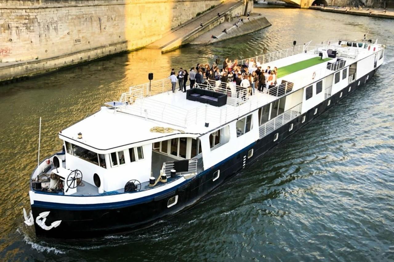 Bateau naviguant sur une rivière avec des passagers debout sur le pont supérieur près d'un quai en pierre.