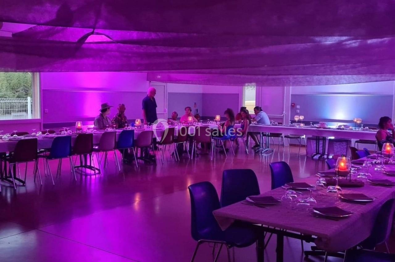 Salle décorée avec des tables dressées et éclairage violet, des convives assis et debout discutant.