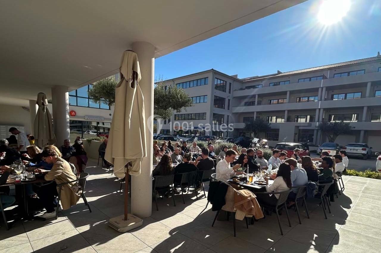 Des personnes déjeunent en terrasse sous un soleil éclatant, entourées de bâtiments modernes et d'arbres.