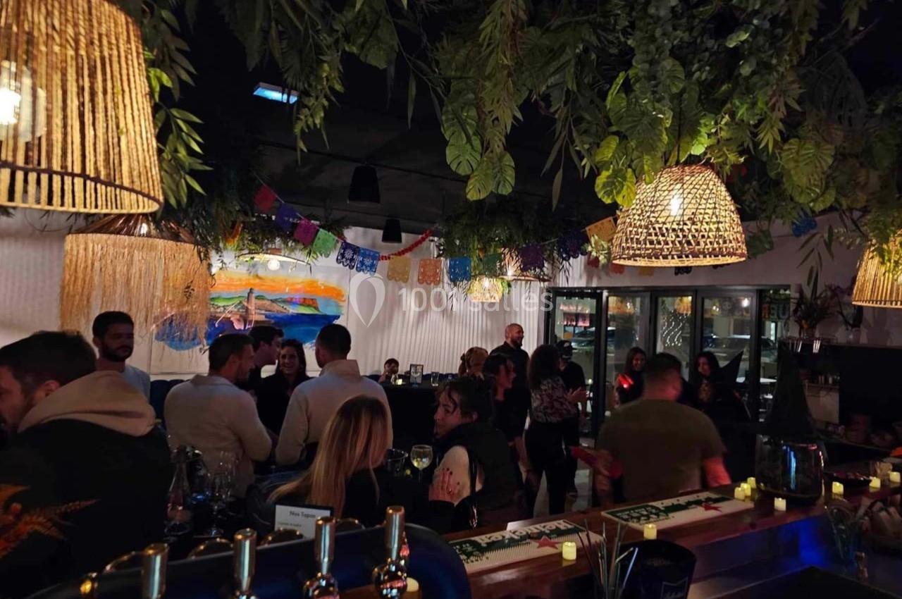 Un bar animé avec des clients discutant sous des lampes suspendues et une décoration végétale.