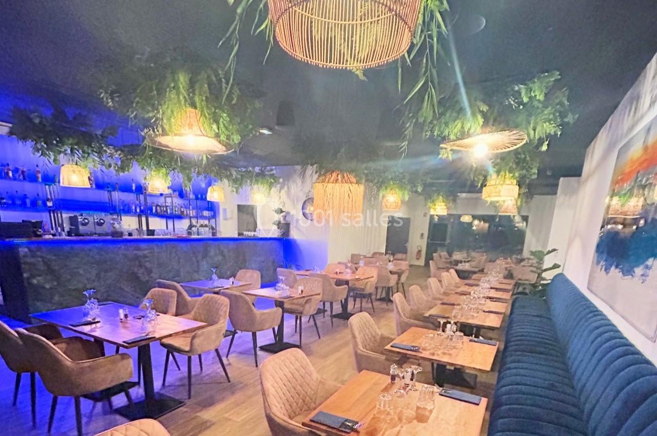 Salle de restaurant moderne avec tables en bois, chaises rembourrées, éclairage tamisé et décoration végétale suspendue.