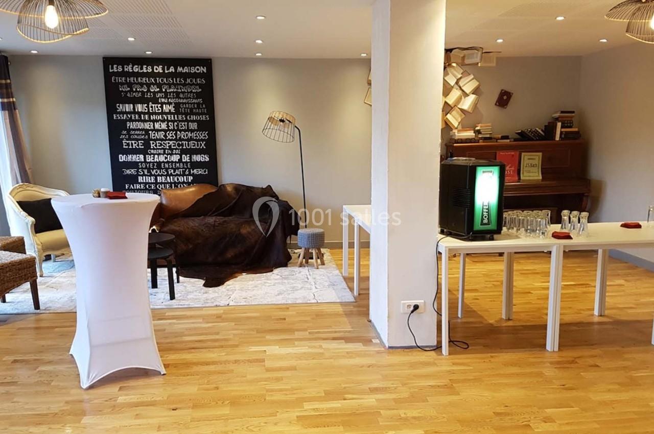 Salle lumineuse avec parquet, mobilier varié, un piano, des tables dressées et une décoration murale textuelle.