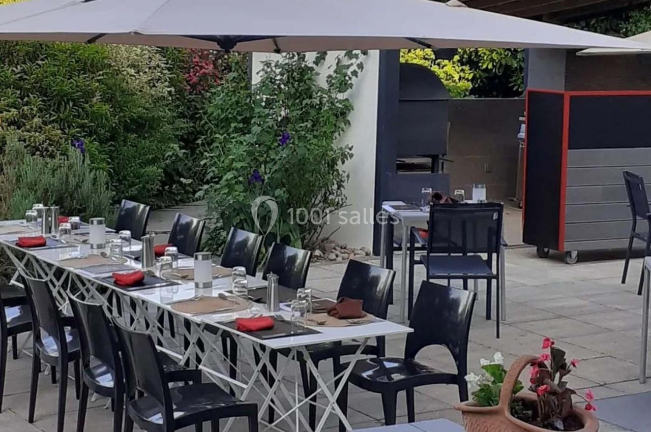 Terrasse aménagée avec une grande table dressée, chaises noires, parasols et un barbecue en arrière-plan.