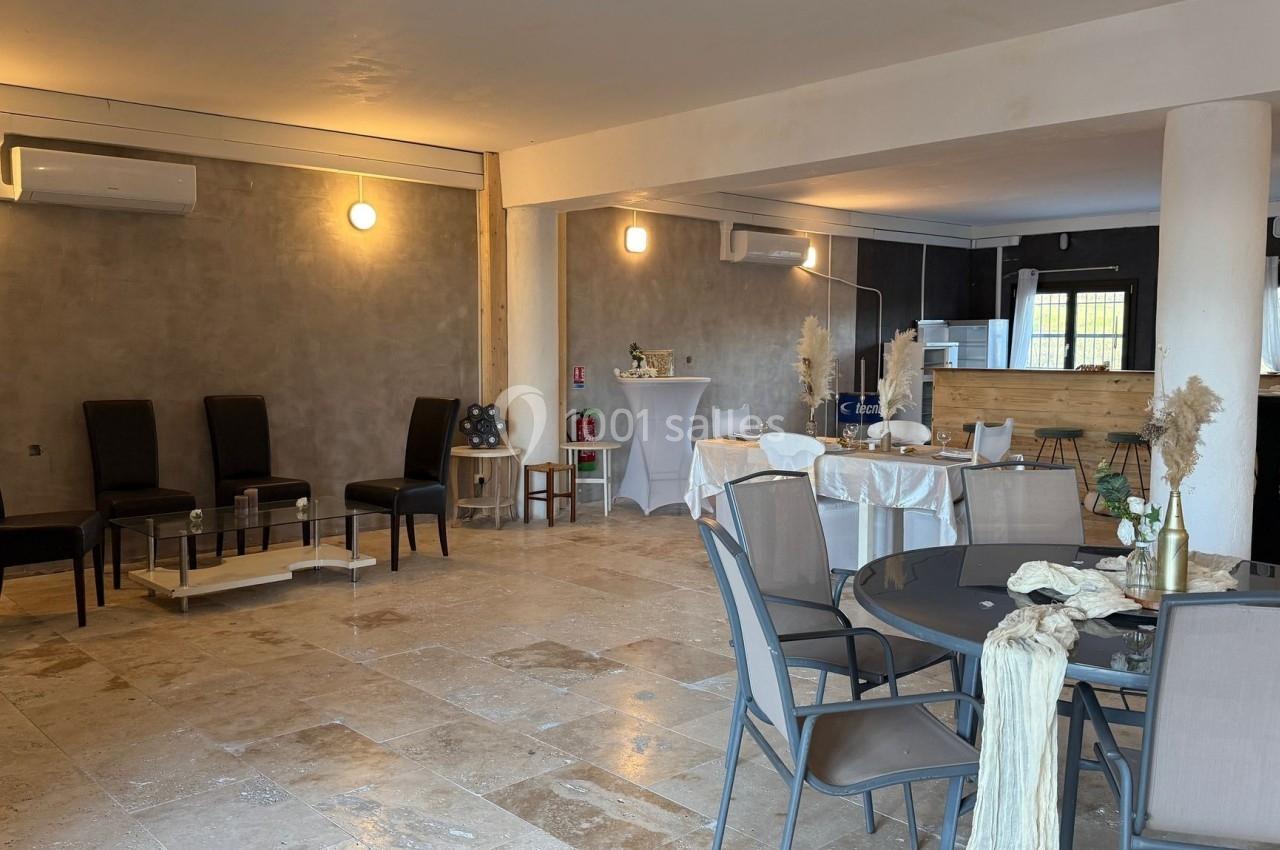 Salle lumineuse avec tables et chaises, décorée pour un événement, avec un coin cuisine visible en arrière-plan.