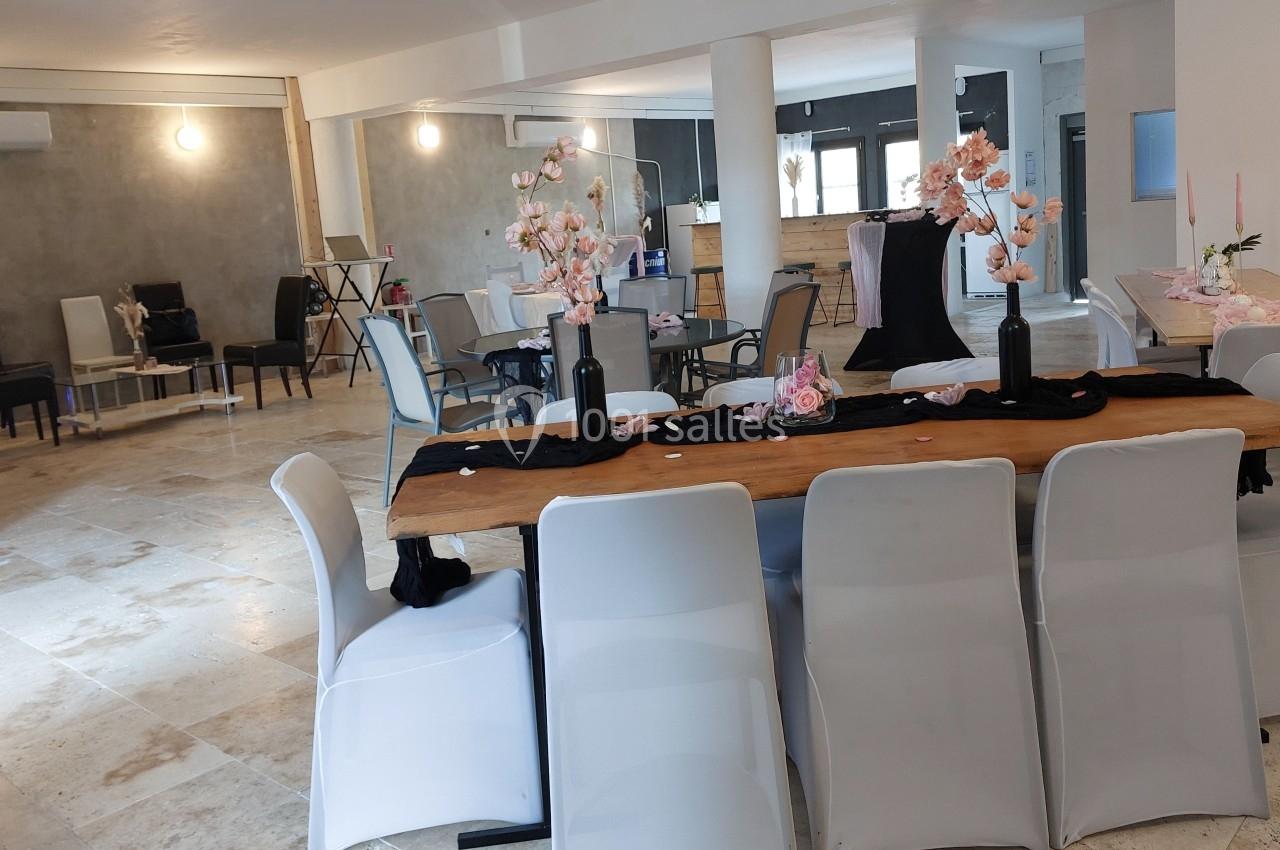 Salle lumineuse avec une grande table décorée de fleurs roses, entourée de chaises blanches, et un espace en arrière-plan.