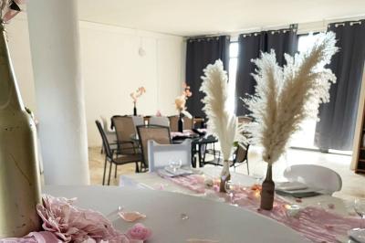 Salle lumineuse avec une grande table décorée de fleurs roses, entourée de chaises blanches, et un espace en arrière-plan.