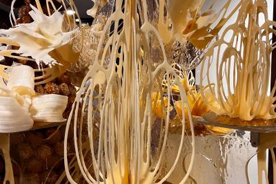 Pièce montée en choux décorée de fleurs en sucre et de motifs en chocolat blanc, posée devant un mur de briques.