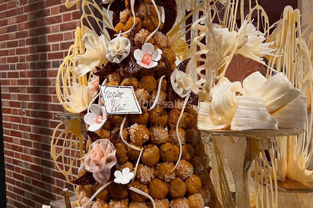Pièce montée en choux décorée de fleurs en sucre et de motifs en chocolat blanc, posée devant un mur de briques.