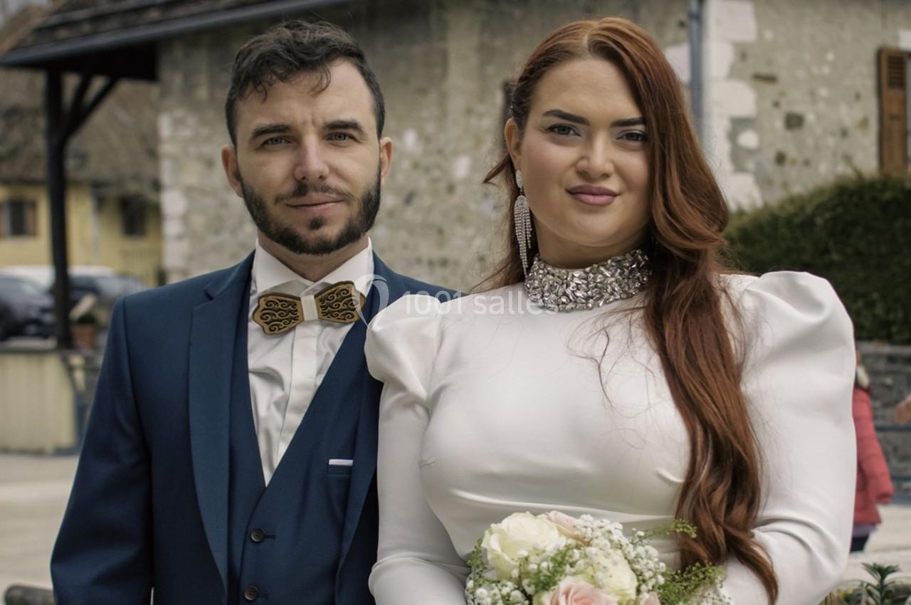 Un couple en tenue de mariage pose devant un bâtiment en pierre dans un cadre extérieur.