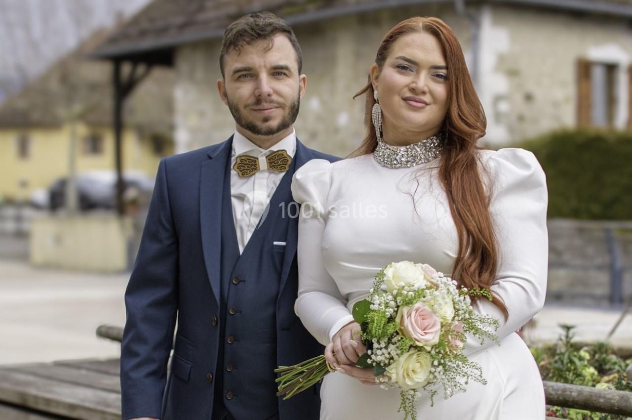 Un couple en tenue de mariage pose dehors, la mariée tenant un bouquet de fleurs.