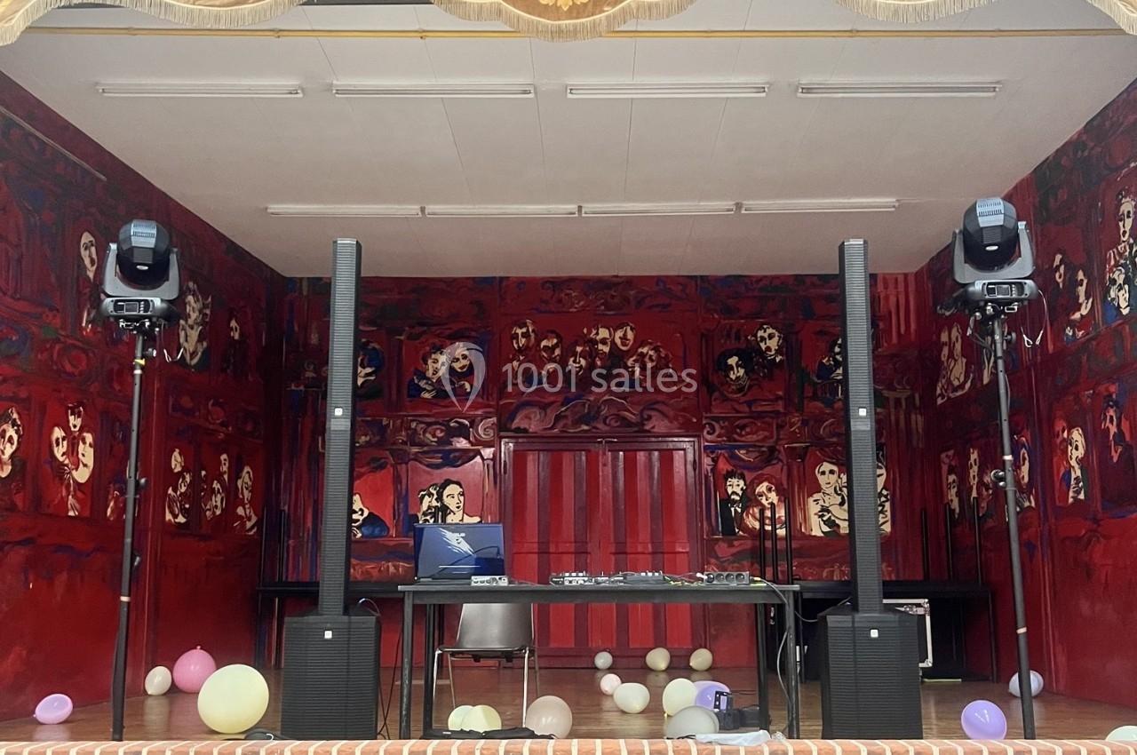 Scène décorée de peintures rouges avec des enceintes, une table équipée de matériel audio et des ballons au sol.