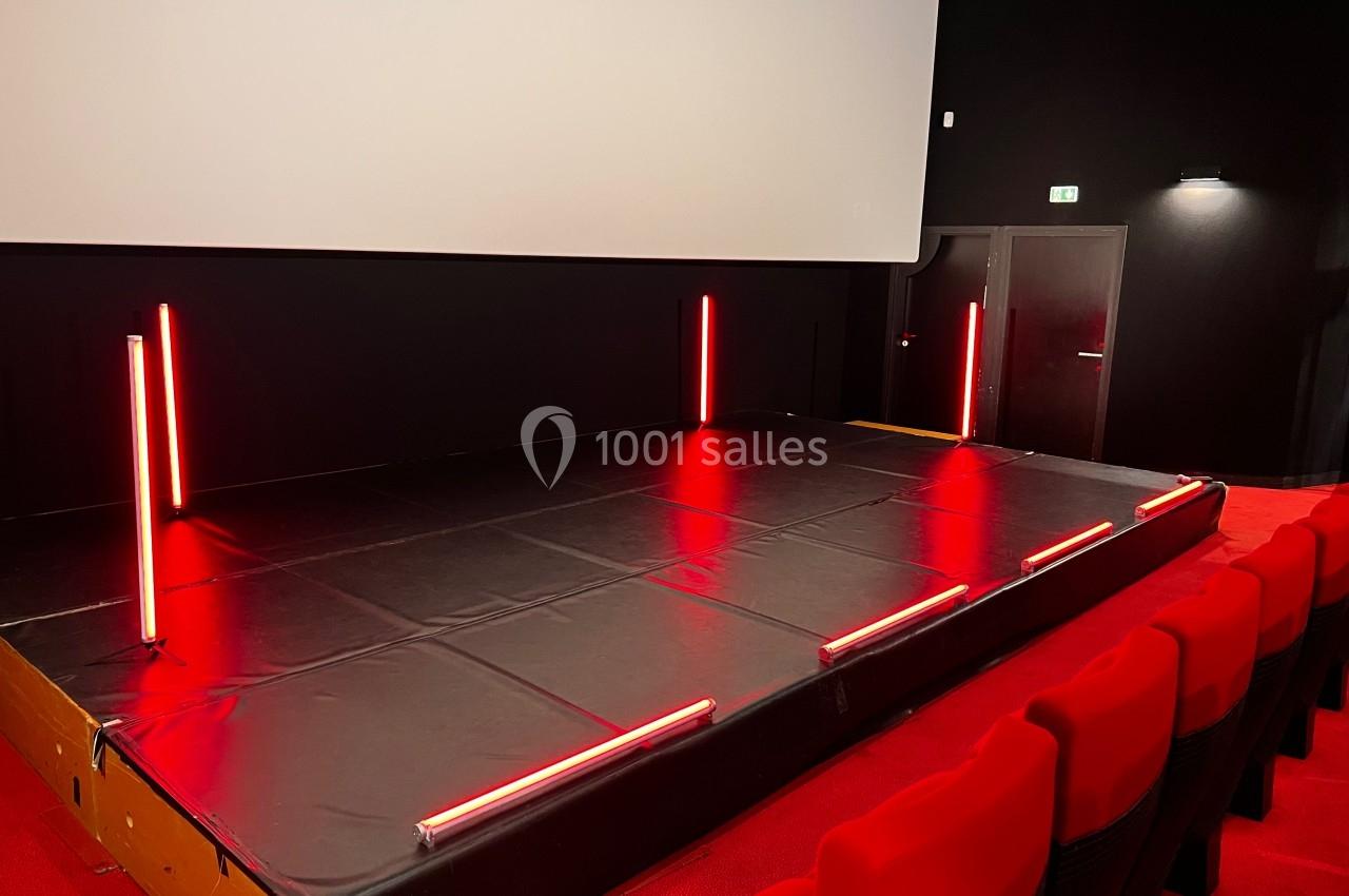 Scène surélevée avec éclairage rouge néon, située dans une salle de spectacle aux sièges rouges et écran blanc.
