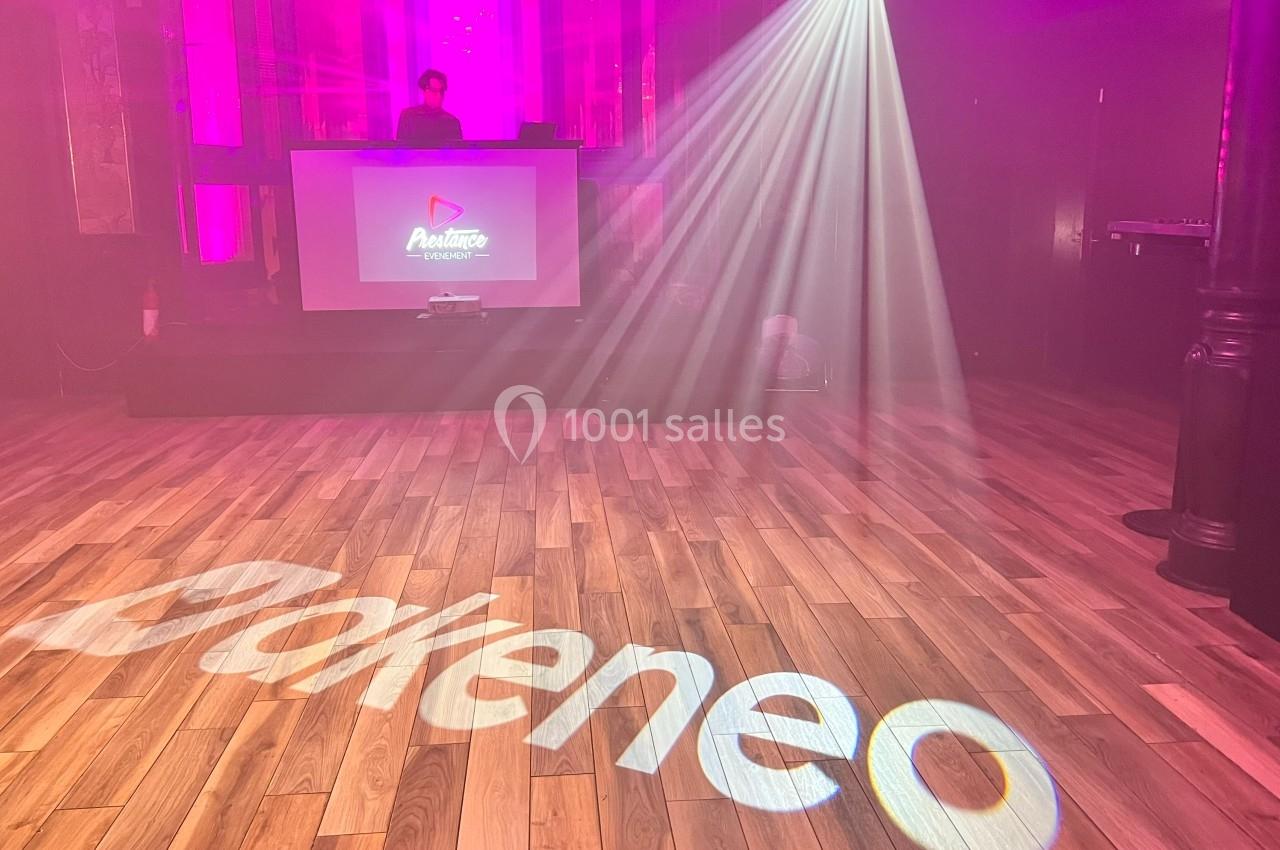 Salle de spectacle avec éclairage coloré, logo ’Akeneo’ projeté au sol et un DJ sur une scène en arrière-plan.