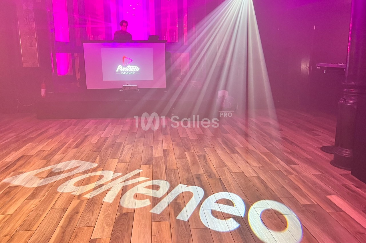 Salle de spectacle avec éclairage coloré, logo ’Akeneo’ projeté au sol et un DJ sur une scène en arrière-plan.