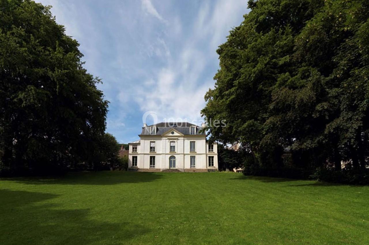 Manoir blanc entouré d'arbres, avec une pelouse verte au premier plan et un ciel partiellement nuageux.