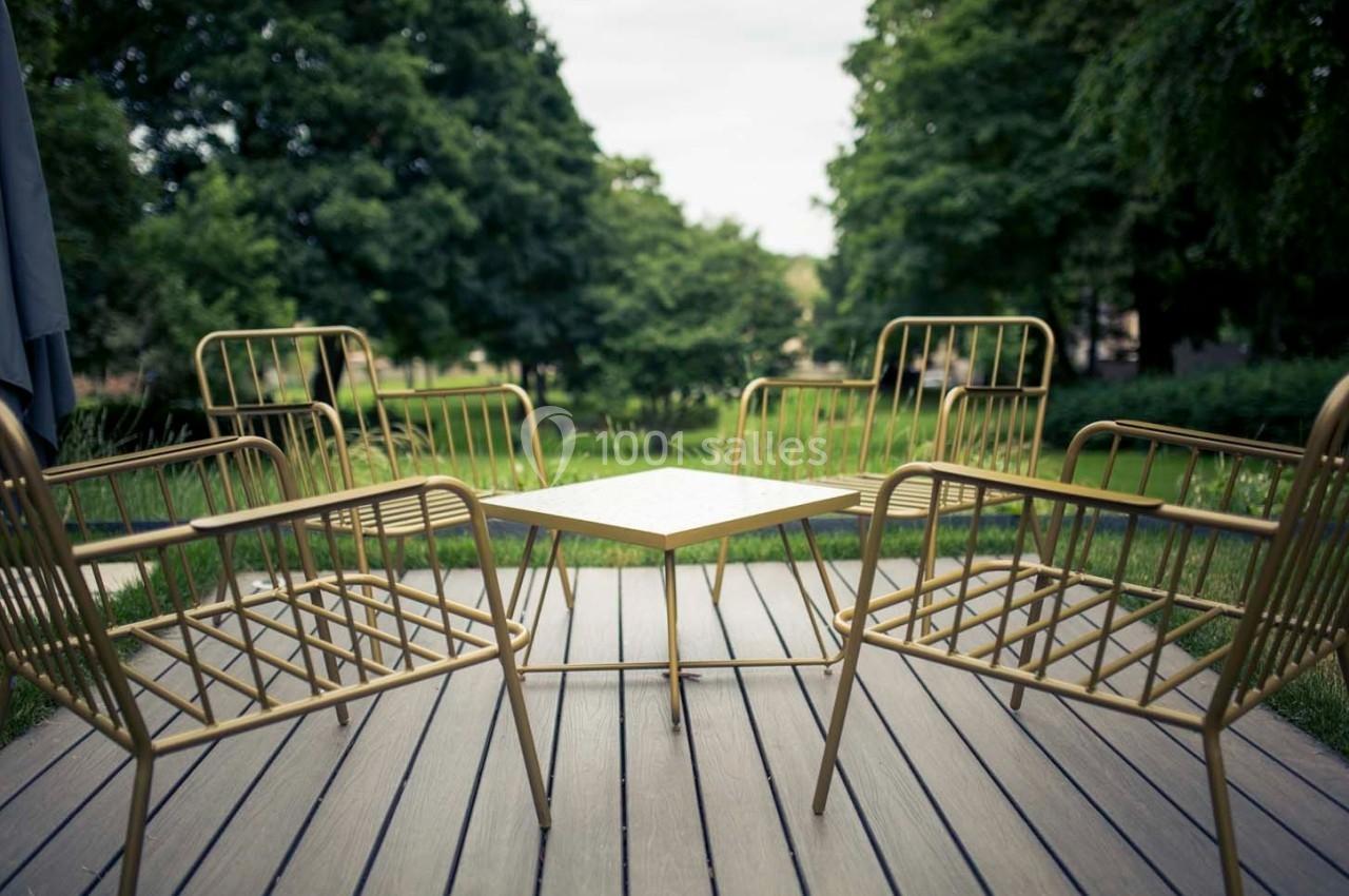 Quatre chaises en métal doré entourent une table carrée sur une terrasse en bois, avec un jardin verdoyant en arrière-plan.