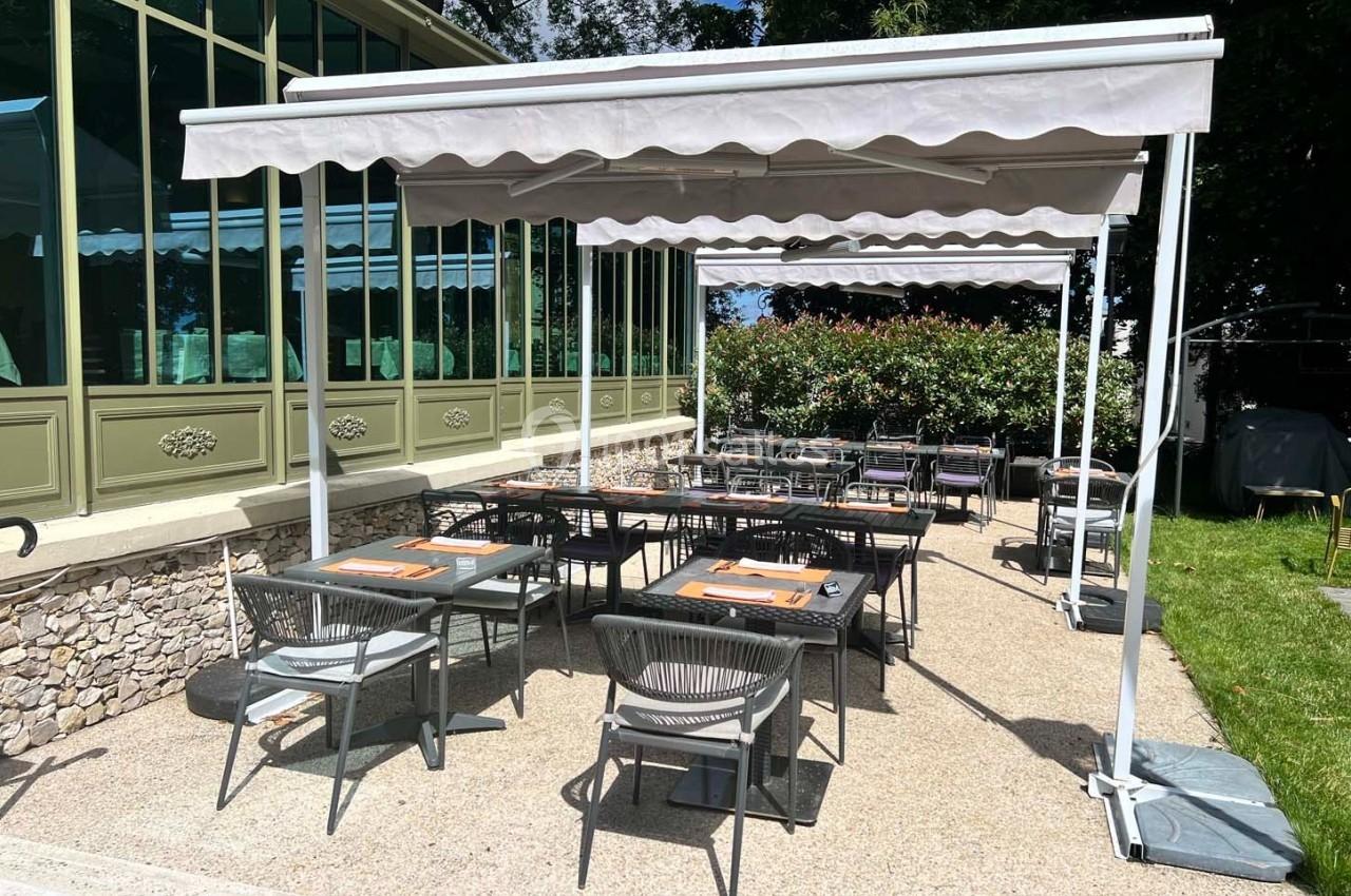Terrasse extérieure d'un restaurant avec tables dressées, chaises en métal et parasols, bordée de verdure et d'une verrière.