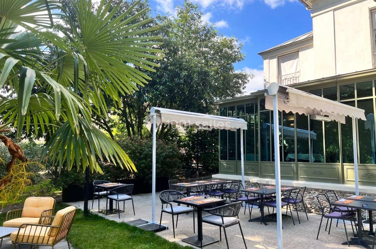 Terrasse extérieure avec tables et chaises, entourée de végétation et attenante à un bâtiment avec grandes fenêtres.