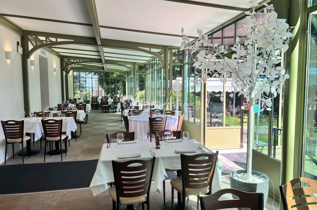 Salle de restaurant lumineuse avec grandes baies vitrées, tables dressées et vue sur un jardin verdoyant.