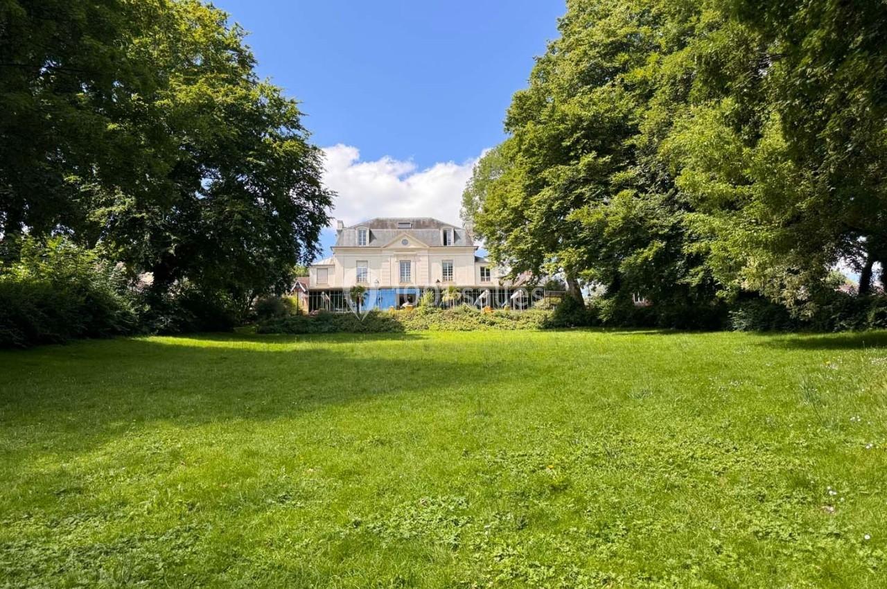 Pelouse verdoyante bordée d'arbres avec une grande maison blanche en arrière-plan sous un ciel bleu.
