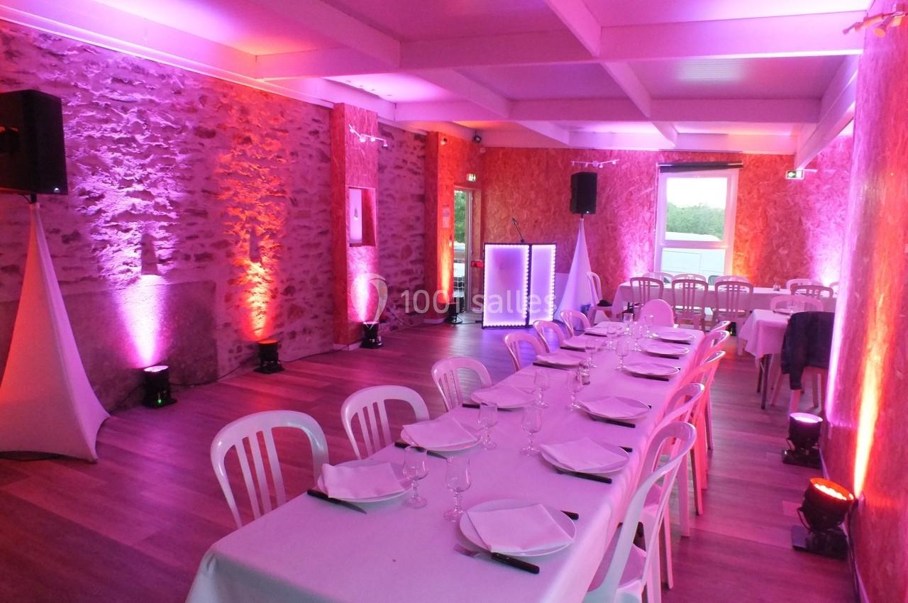 Salle de réception avec tables dressées, chaises blanches et éclairage d'ambiance rose et violet.
