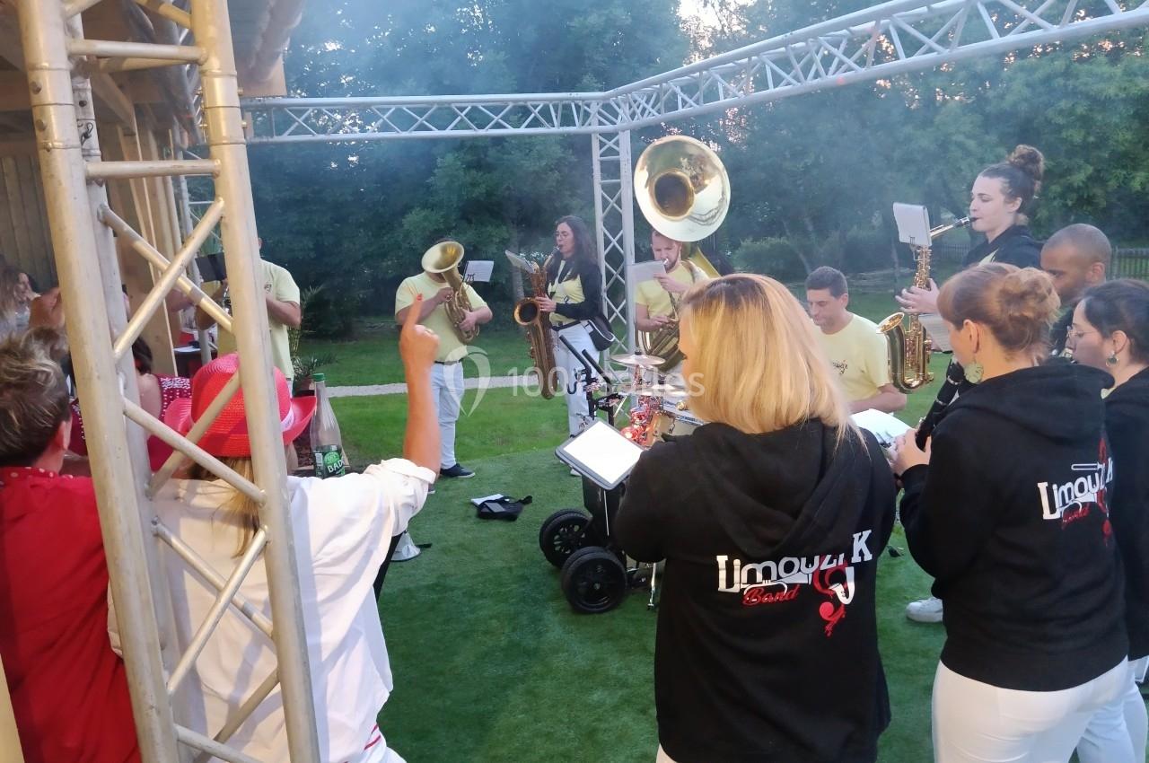 Un groupe de musiciens en plein air joue des instruments à vent et à percussion sous une structure métallique.
