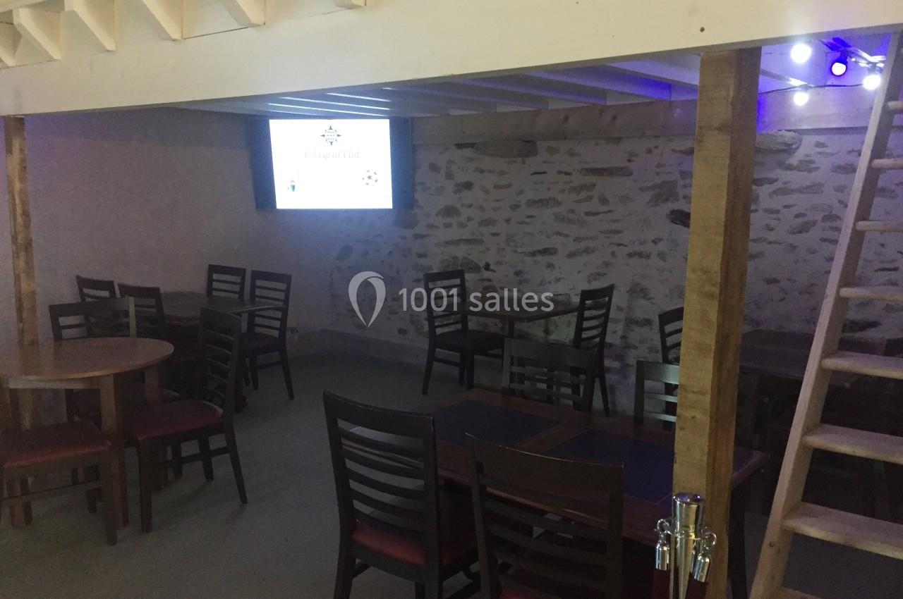 Salle avec des tables et chaises en bois, éclairage tamisé, mur en pierre et écran allumé au fond.