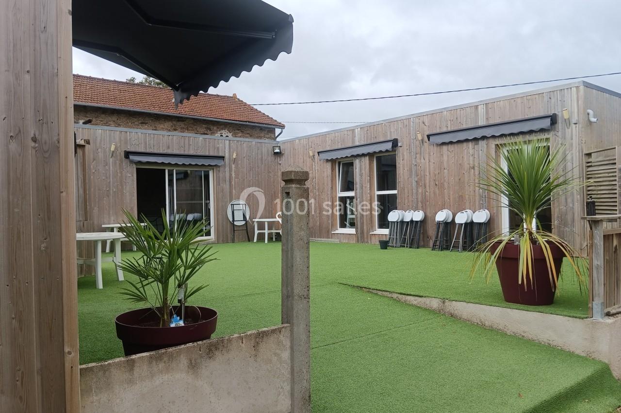 Cour extérieure avec gazon synthétique, tables, chaises empilées, plantes en pot et bâtiments en bois autour.