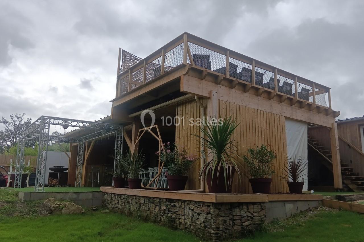 Structure en bois à deux niveaux avec terrasse supérieure, plantes en pots et pelouse environnante sous un ciel nuageux.