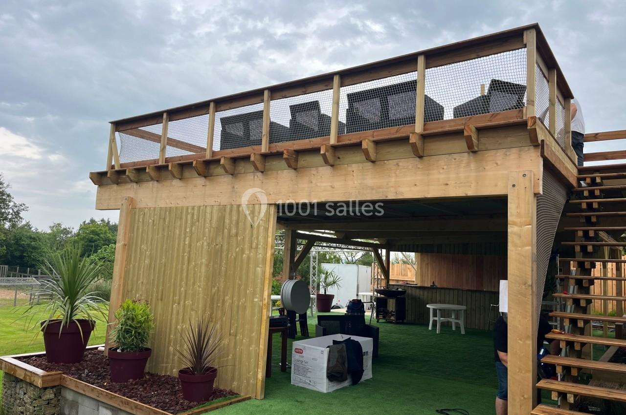 Structure en bois à deux niveaux avec terrasse aménagée en haut et espace couvert en bas, entourée de végétation.