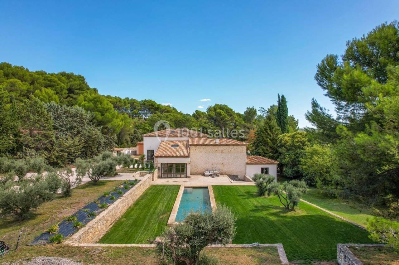 Maison en pierre avec piscine centrale, entourée d'un jardin verdoyant et d'arbres, sous un ciel dégagé.