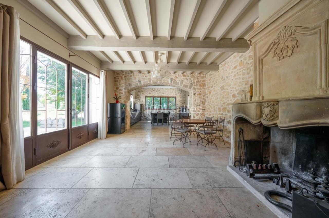 Salle lumineuse avec cheminée en pierre, poutres apparentes, sol en pierre et grandes fenêtres donnant sur un jardin.