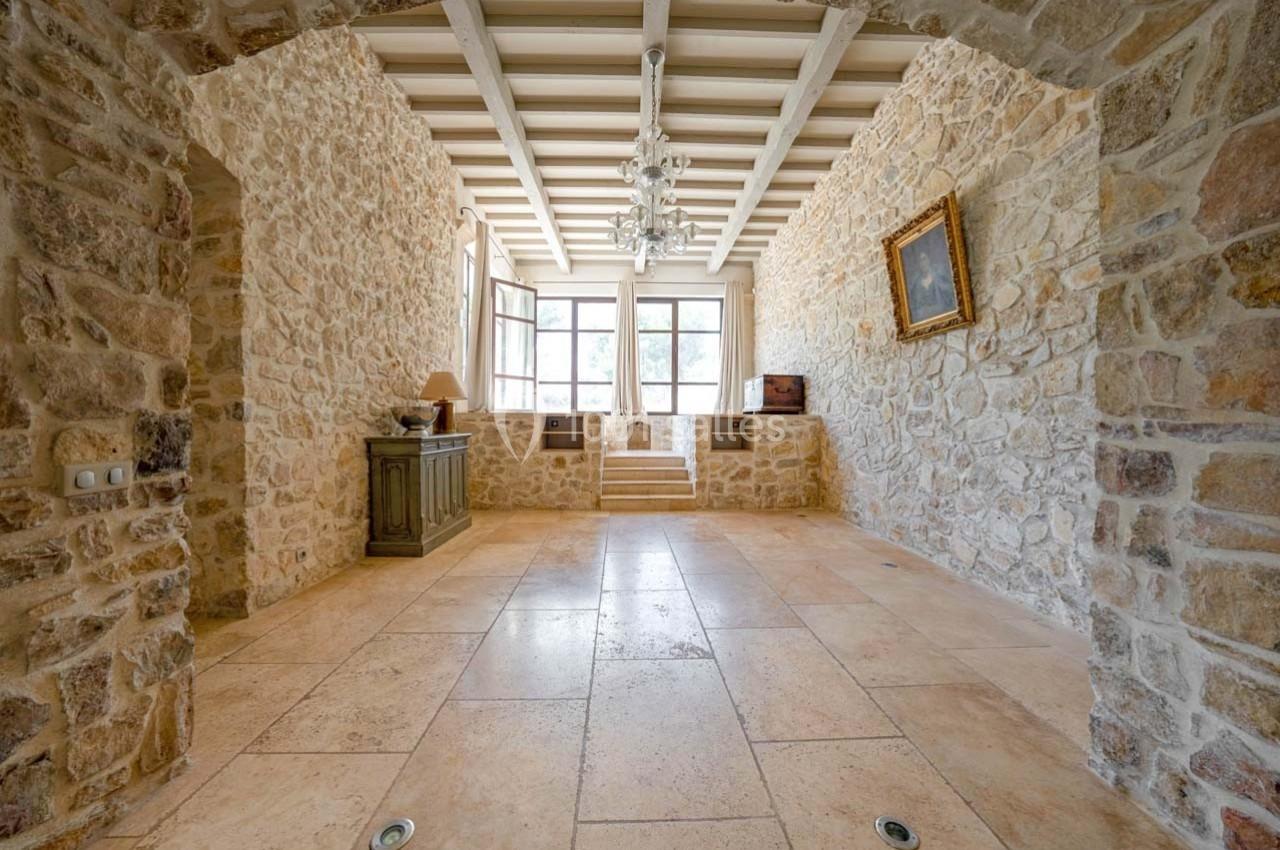 Salle lumineuse avec murs en pierre, sol en carreaux beige, poutres apparentes et fenêtres donnant sur l'extérieur.