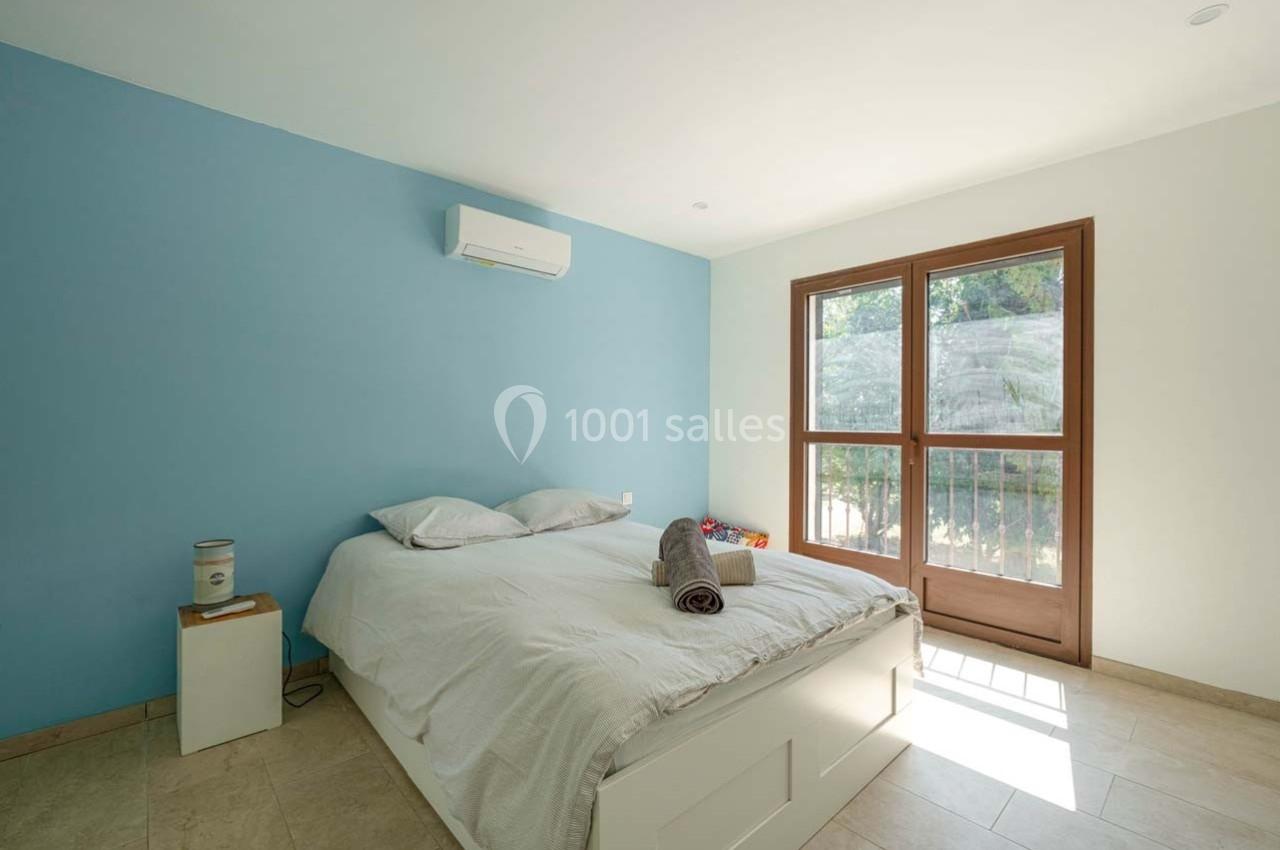 Chambre lumineuse avec un mur bleu, un lit double, une fenêtre donnant sur un jardin et un climatiseur mural.