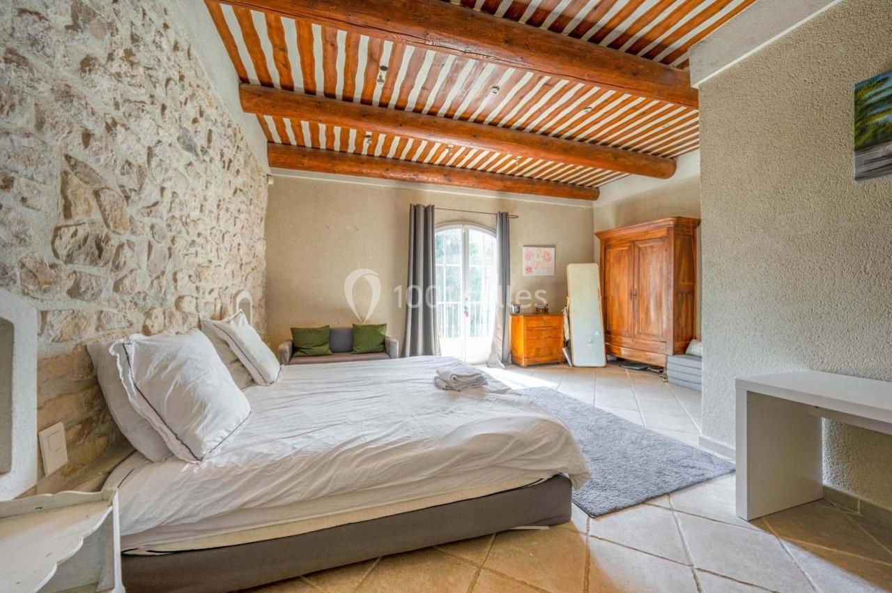 Chambre lumineuse avec lit double, murs en pierre, poutres apparentes, armoire en bois et grande fenêtre donnant sur l…