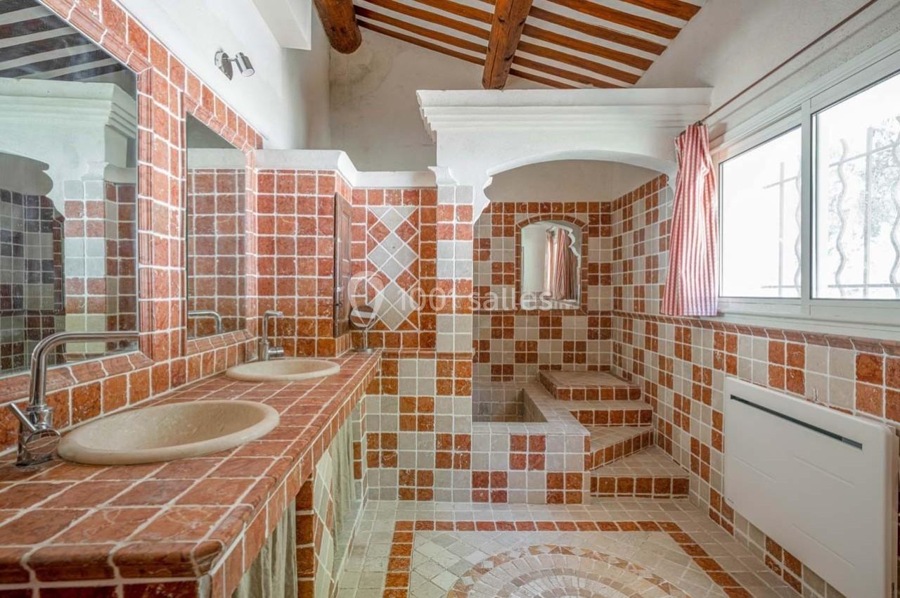 Salle de bain avec carrelage en terre cuite, double vasque, douche à l'italienne et plafond avec poutres apparentes.
