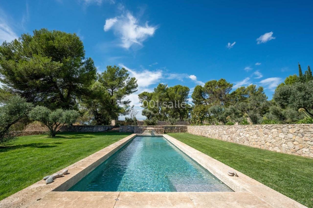 Piscine rectangulaire entourée de pelouse, d'arbres et de murs en pierre sous un ciel bleu dégagé.