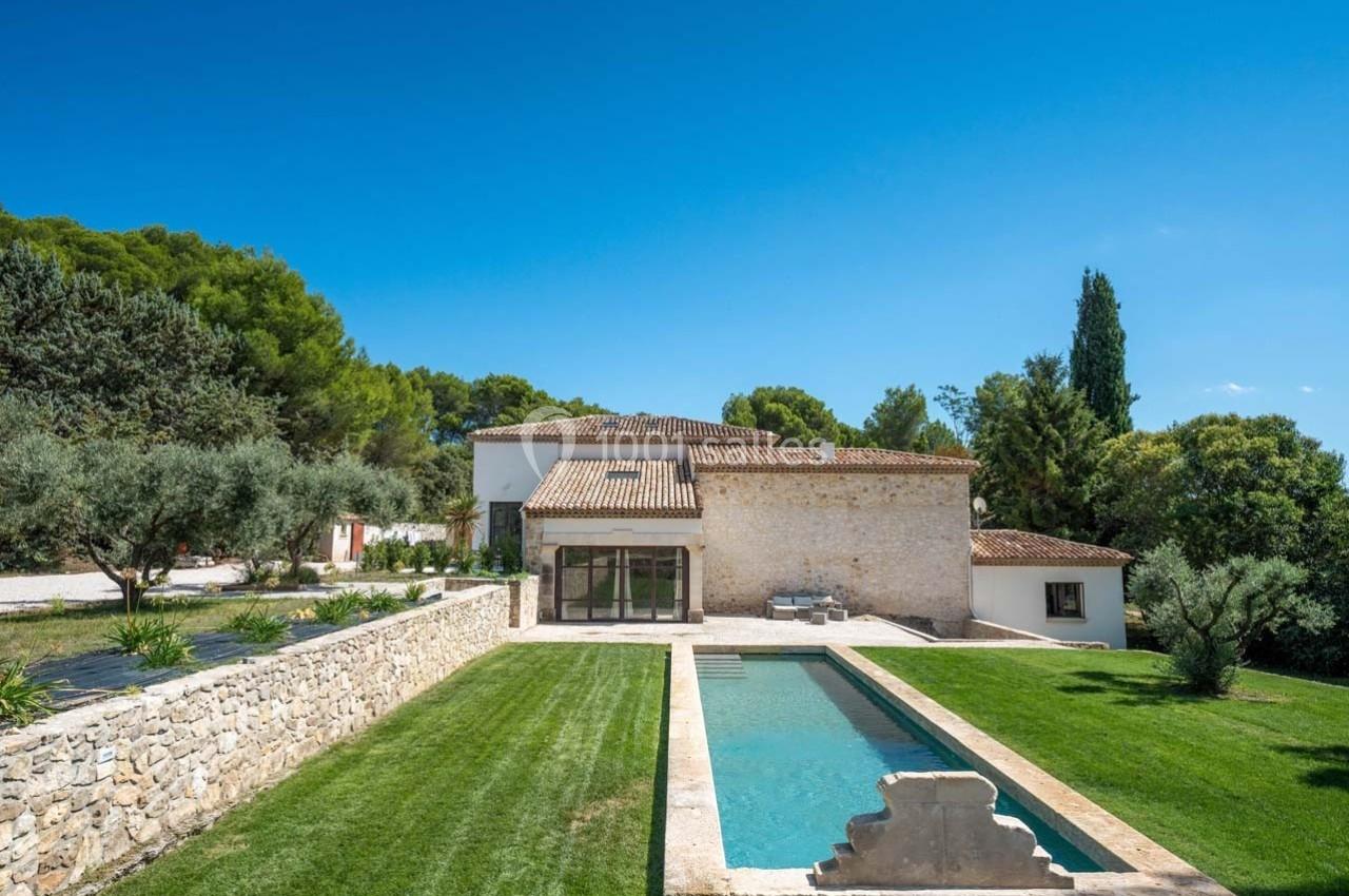 Maison en pierre avec piscine rectangulaire, entourée de pelouses et d'arbres, sous un ciel bleu clair.