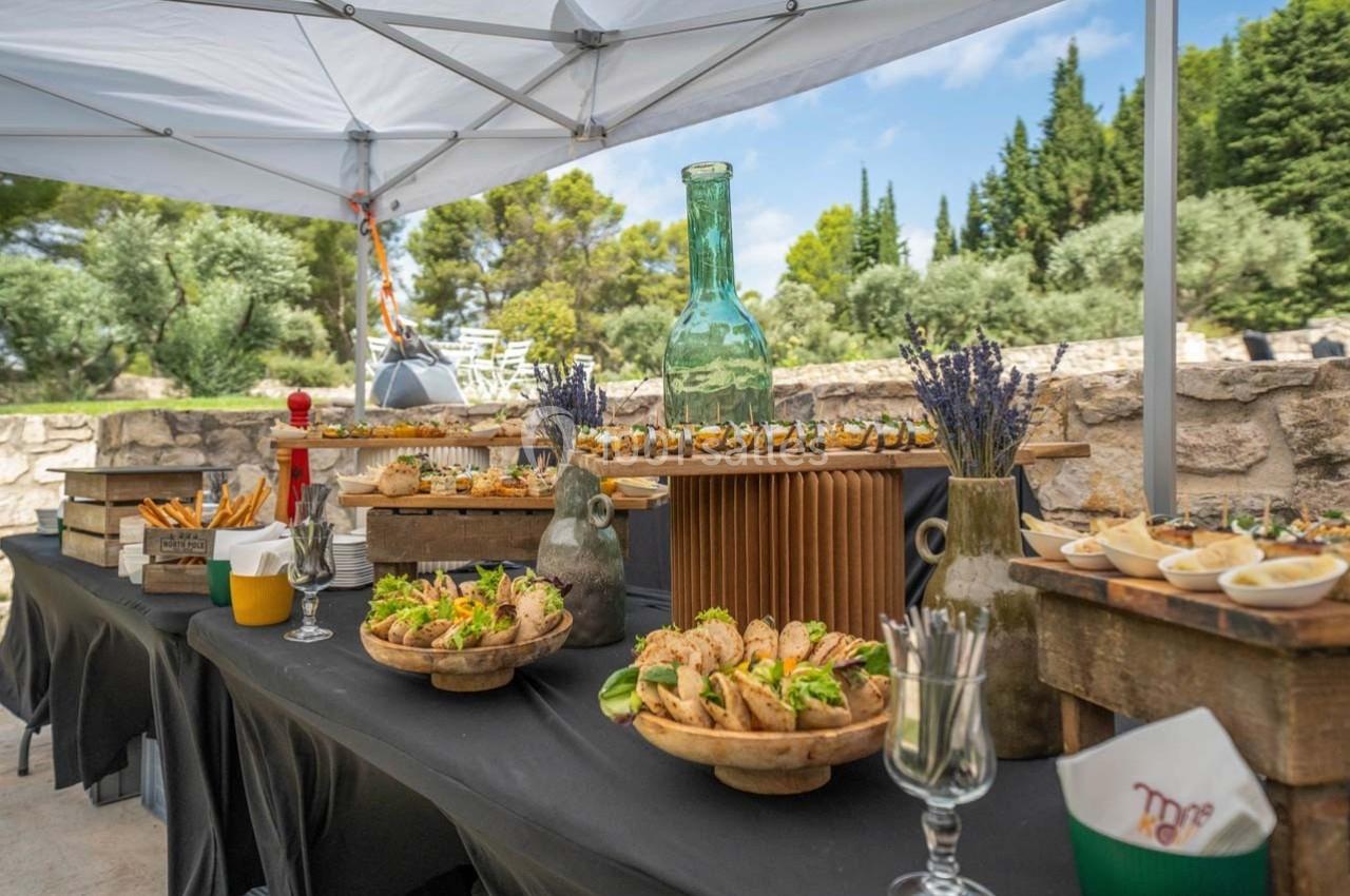 Buffet en plein air avec divers plats et boissons disposés sur des tables sous un auvent, entouré de verdure.