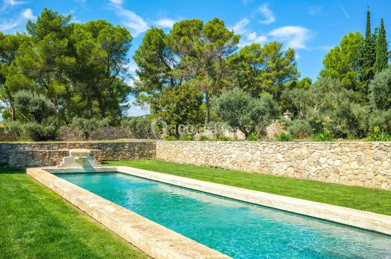 Piscine rectangulaire en pierre entourée de pelouse, avec des arbres et un mur en pierre en arrière-plan.