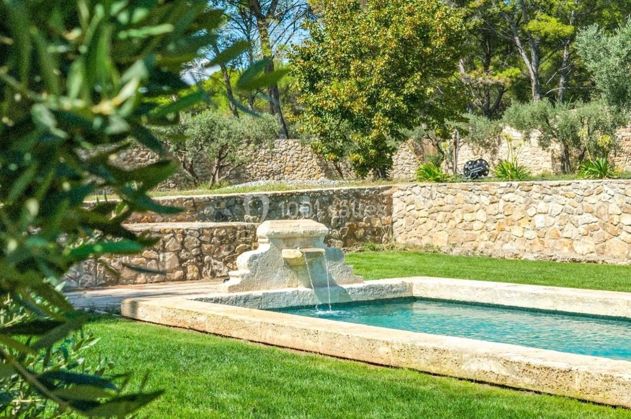 Bassin rectangulaire en pierre avec une fontaine, entouré de pelouse, d'arbres et d'un mur en pierre.