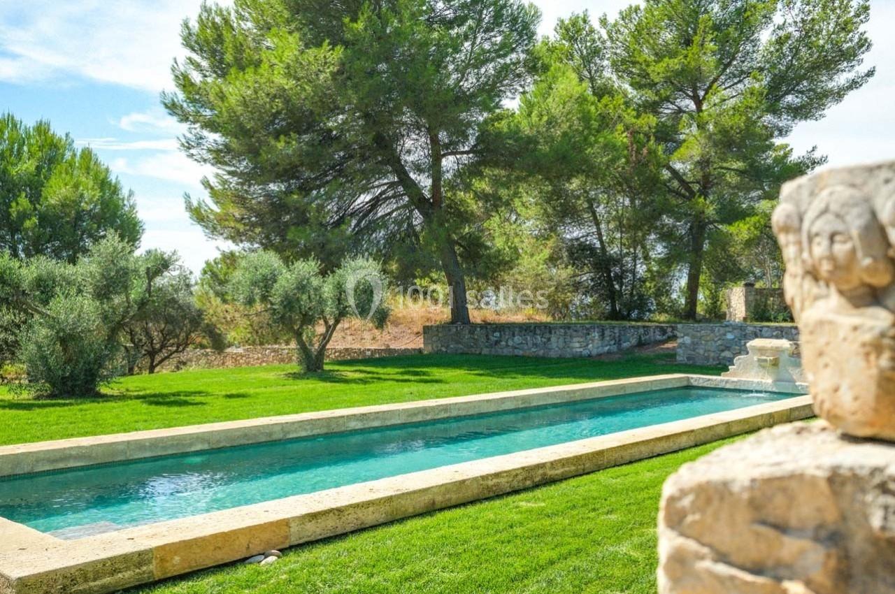 Piscine rectangulaire entourée de pelouse, d'oliviers et de pins, avec un mur en pierre au second plan.