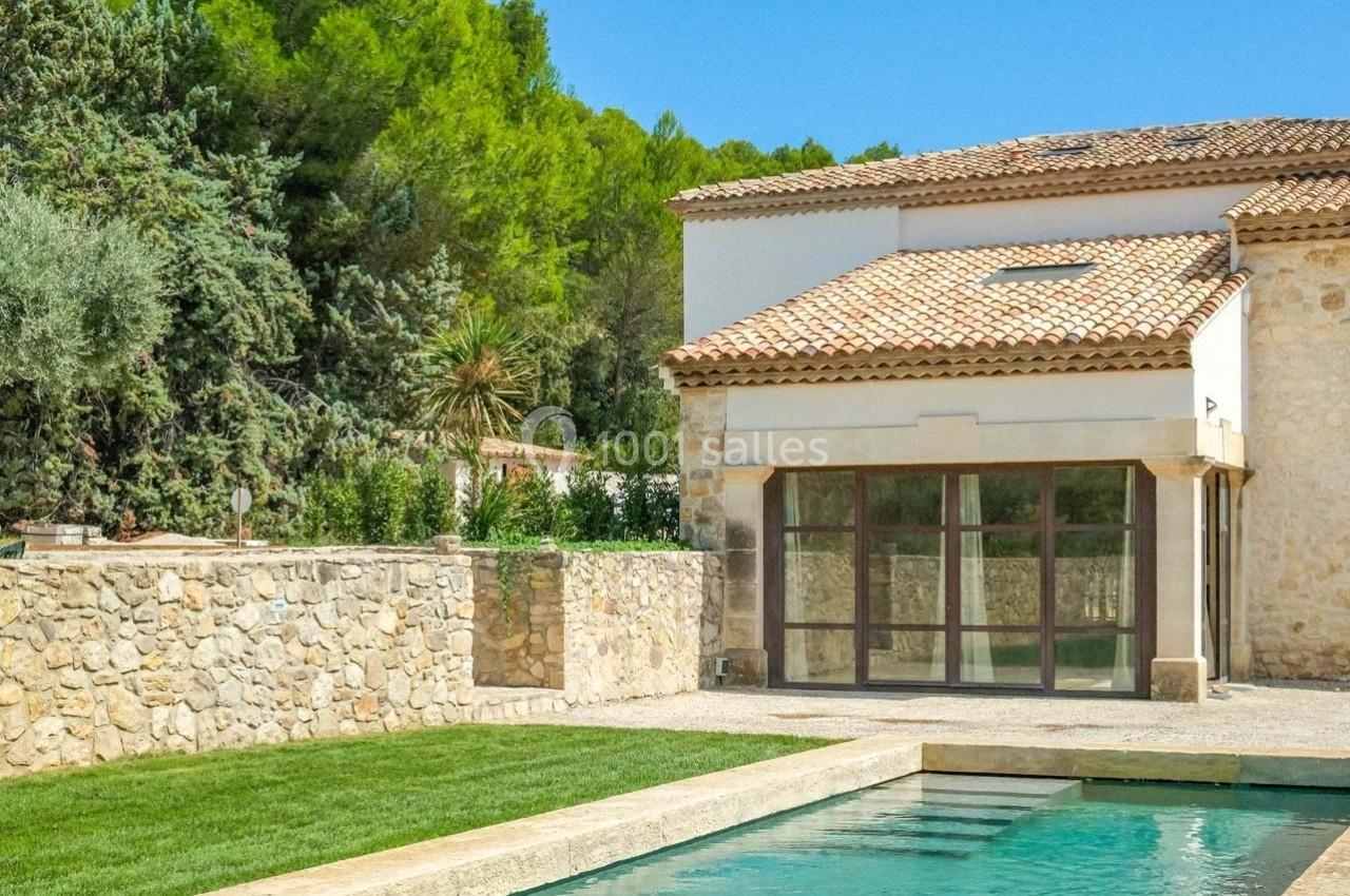 Maison en pierre avec toit en tuiles, grande baie vitrée, jardin arboré et piscine au premier plan.