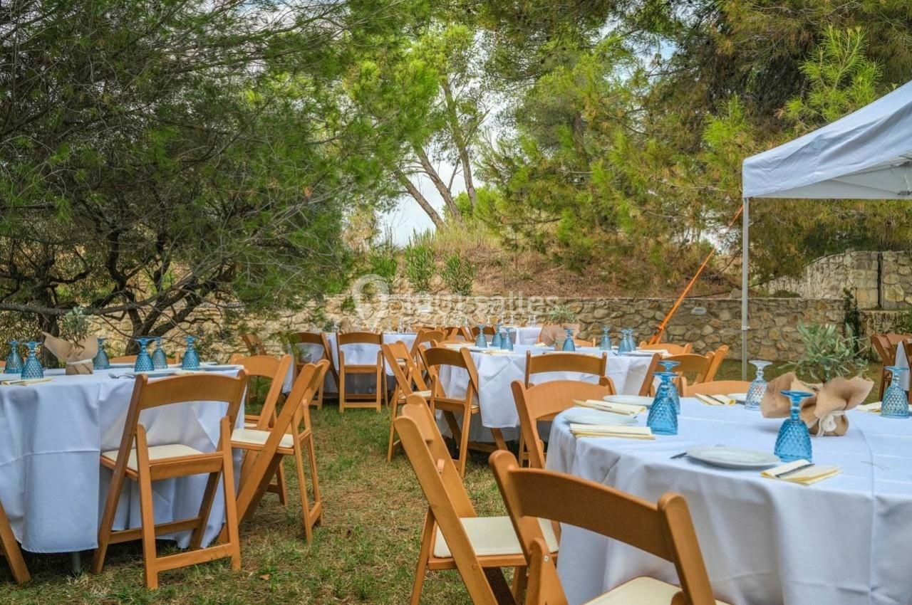 Tables dressées avec nappes blanches, chaises en bois et décorations, installées en extérieur sous des arbres.