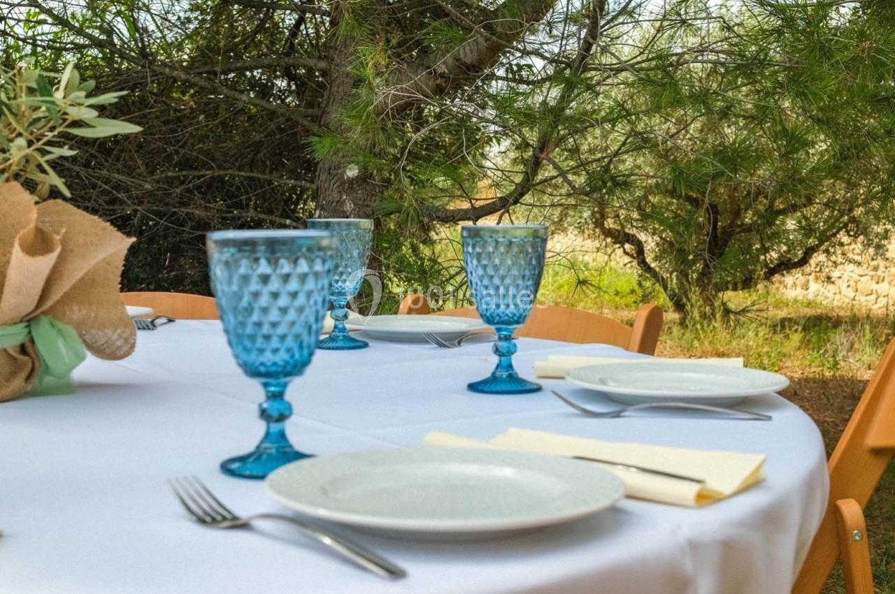 Table dressée en extérieur avec nappes blanches, verres bleus, assiettes blanches et couverts, entourée de végétation.