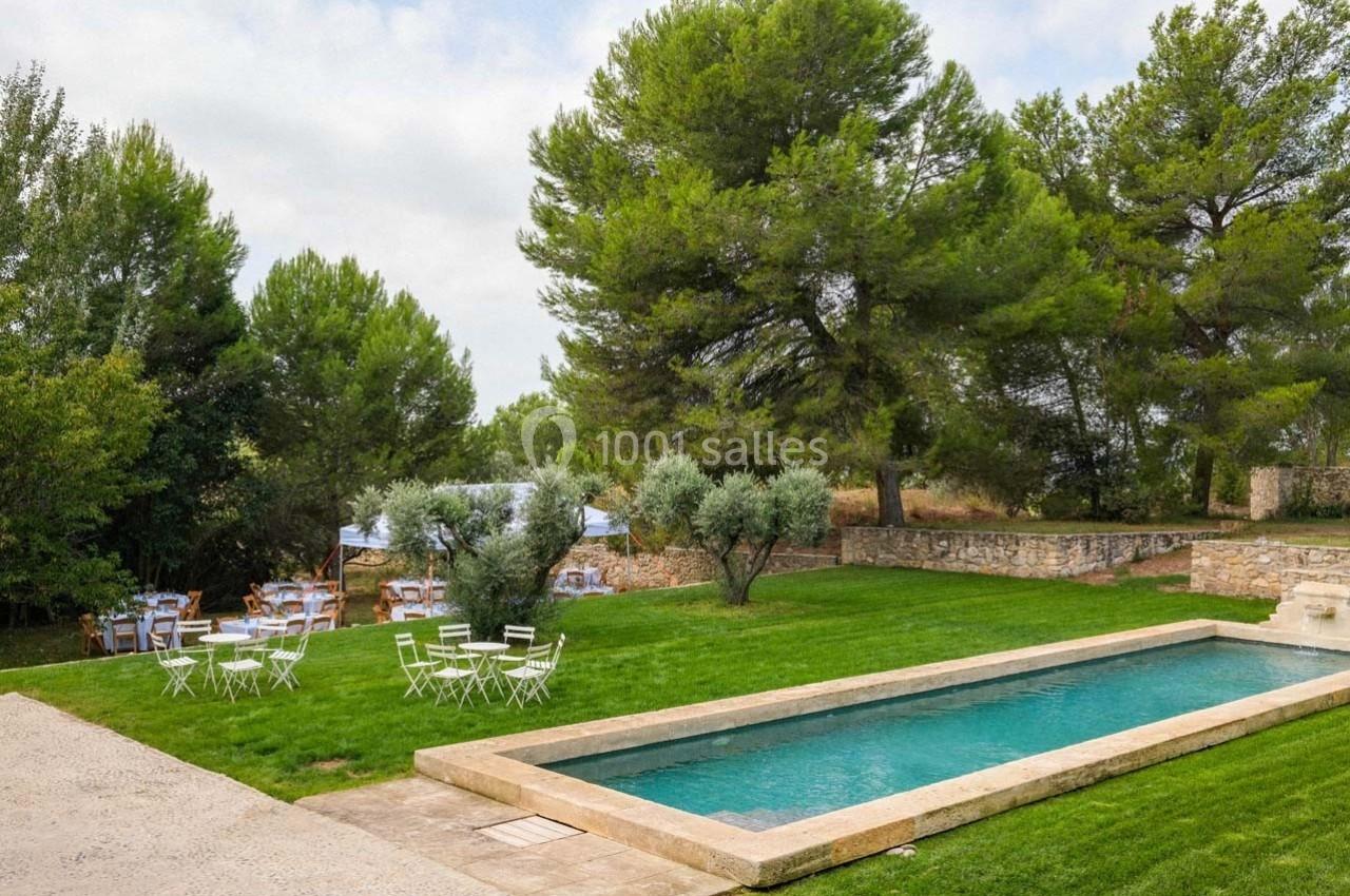 Jardin avec piscine rectangulaire, tables et chaises blanches sur pelouse, entouré d'arbres et d'un mur en pierre.