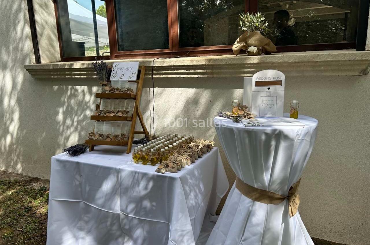 Table décorée pour un événement, avec flacons d'huile d'olive, sachets cadeaux et pancartes sur des nappes blanches.