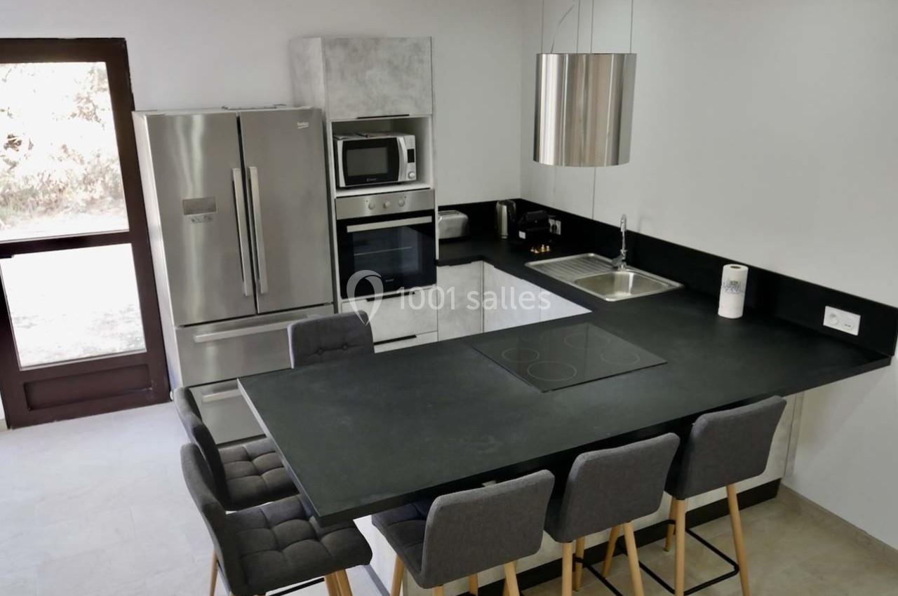 Cuisine moderne avec îlot central noir, tabourets gris, électroménagers en inox et évier sous une hotte suspendue.