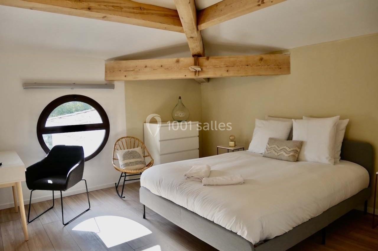 Chambre lumineuse avec lit double, poutres apparentes, bureau, chaise, commode et fenêtre ronde.