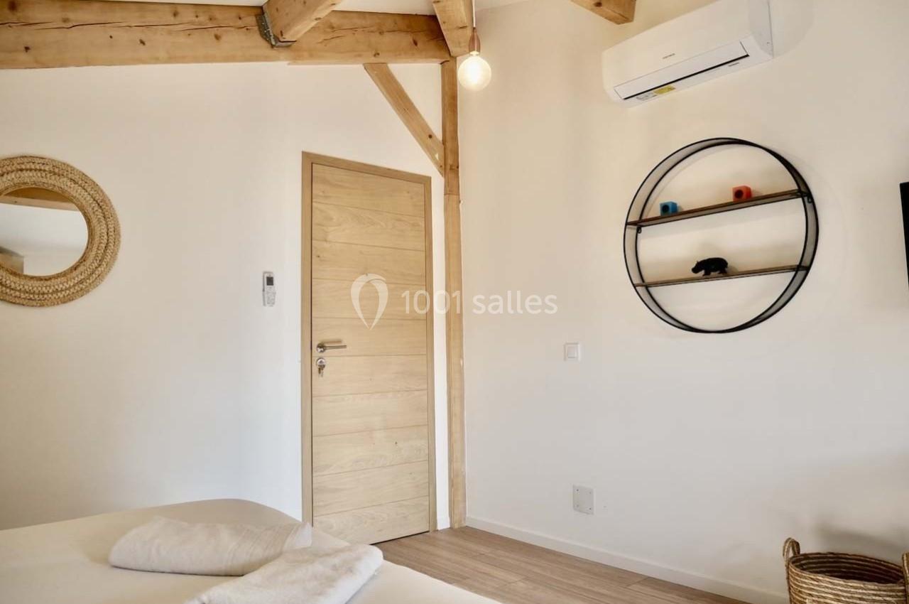 Chambre lumineuse avec poutres apparentes, lit, porte en bois, miroir rond, étagère murale et climatisation.